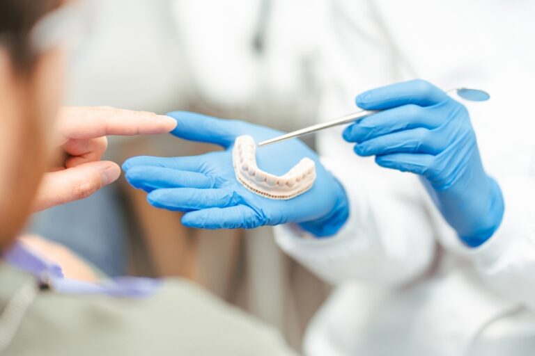 A dentist in blue gloves holds a dental mold and points to it with a metal tool while a patient gestures toward the mold, possibly discussing dental treatment or procedure options.