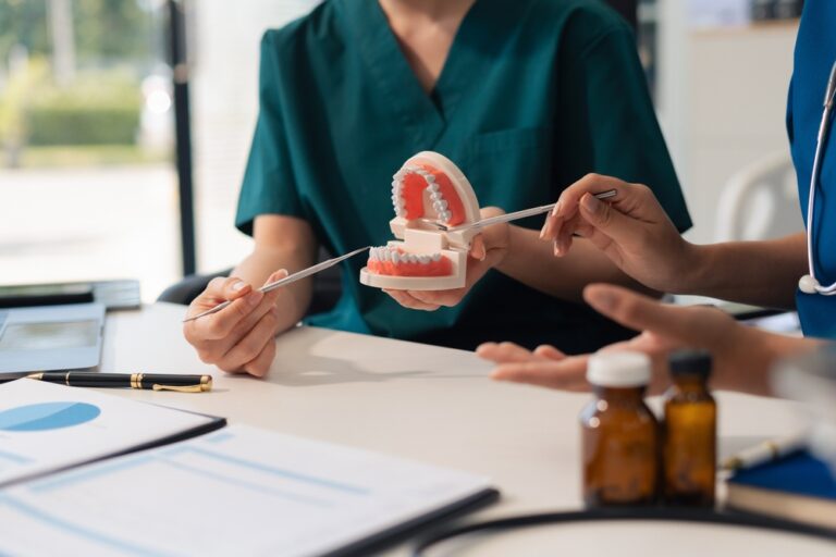Two people in medical scrubs sit at a desk. One holds a dental model and points to it with a dental tool, while the other gestures. Medicine bottles and charts are on the table.