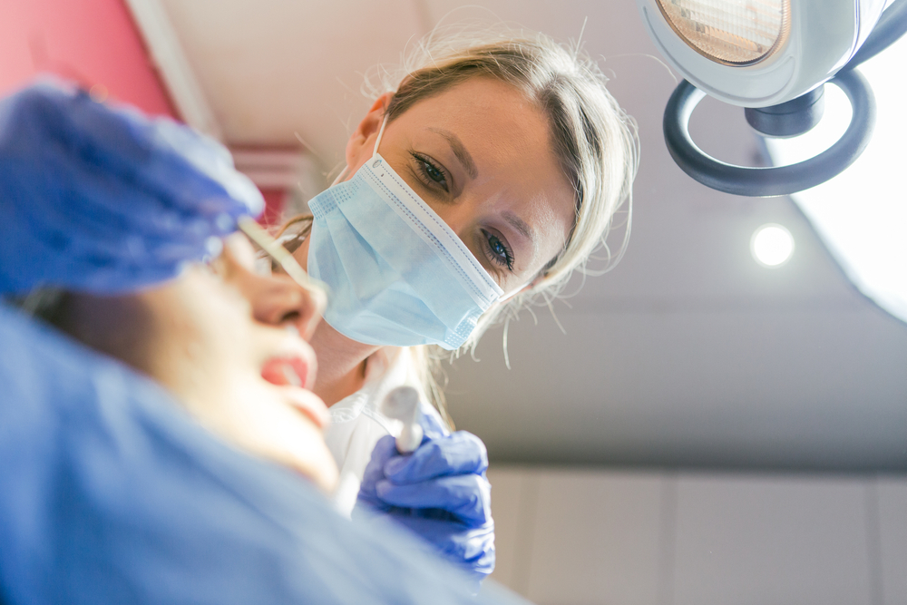 Photo of a dentist explaining the process of implant to a patient using a model of a jaw with a dental implant. The dentist is pointing to the different parts of the implant, while the patient is looking on attentively. No text on image.