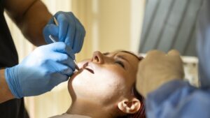 Image of a smiling, healthy middle aged woman, sitting in a dental chair, as a dentist checks her gums. No text on image.